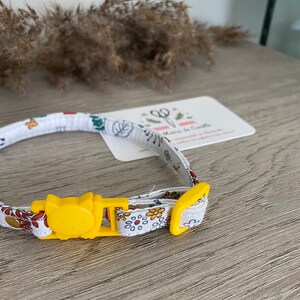 May include: A white cat collar with a yellow buckle and a floral pattern. The buckle has a cat face design.
