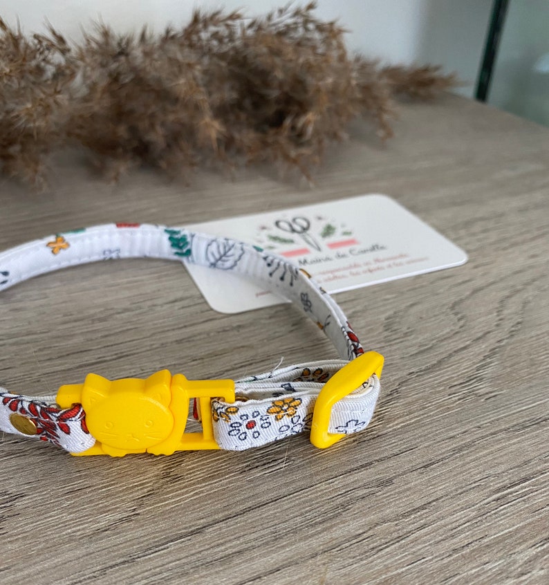 May include: A white cat collar with a yellow buckle and a floral pattern. The buckle has a cat face design.