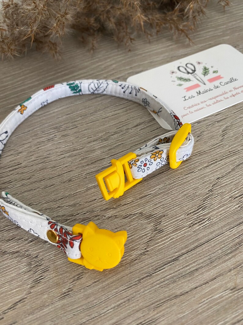 May include: A white and yellow cat collar with a floral pattern and a yellow cat-shaped buckle. The collar is made by Les Mains de Camille, a company that creates eco-responsible products for the home.