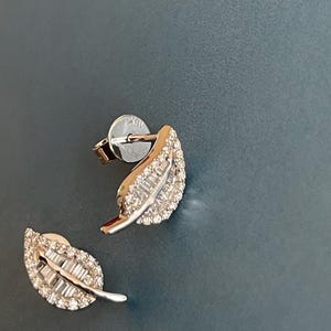 May include: A pair of leaf-shaped stud earrings. The earrings are crafted from silver metal and adorned with small, sparkling gemstones. The leaf design features a central vein and detailed edges, creating a delicate and elegant appearance. The earrings are set against a dark blue background.