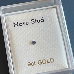 May include: A nose stud with a small, round, purple gemstone, presented in a clear plastic box. The label reads "Nose Stud." The box is set on a white fabric, with "9ct GOLD" printed in gold lettering.