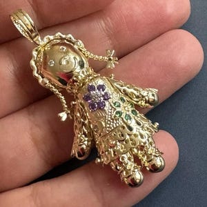 May include: A gold-colored doll pendant with intricate filigree detailing. The doll features a floral design with purple and green gemstones. The pendant is suspended from a loop at the top, ready to be added to a necklace.