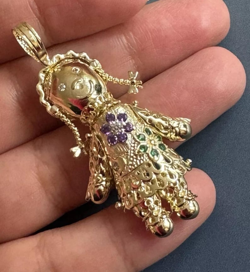 May include: A gold-colored doll pendant with intricate filigree detailing. The doll features a floral design with purple and green gemstones. The pendant is suspended from a loop at the top, ready to be added to a necklace.