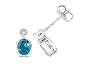 May include: A pair of silver stud earrings. Each earring features an oval, teal-colored gemstone and a small, round, clear gemstone. The earrings are set in a silver metal setting and have a post and butterfly clasp.