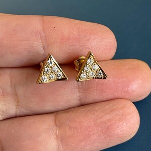 May include: A pair of gold stud earrings, each shaped as a triangle and set with six small, clear, round-cut stones arranged in a triangular pattern. The earrings feature a polished gold metal finish and a secure post backing for attachment.