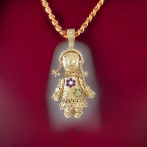 May include: A gold-toned pendant necklace featuring a detailed doll design. The doll has braided hair, a floral design on the dress, and gemstone accents. The necklace chain is a twisted rope style, and the background is a deep red.