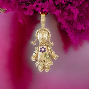May include: A gold-colored pendant in the shape of a doll, with a smiling face, braided pigtails, and a floral design on the dress. The doll pendant has a loop at the top for a chain. The background is a gradient of pink and magenta.