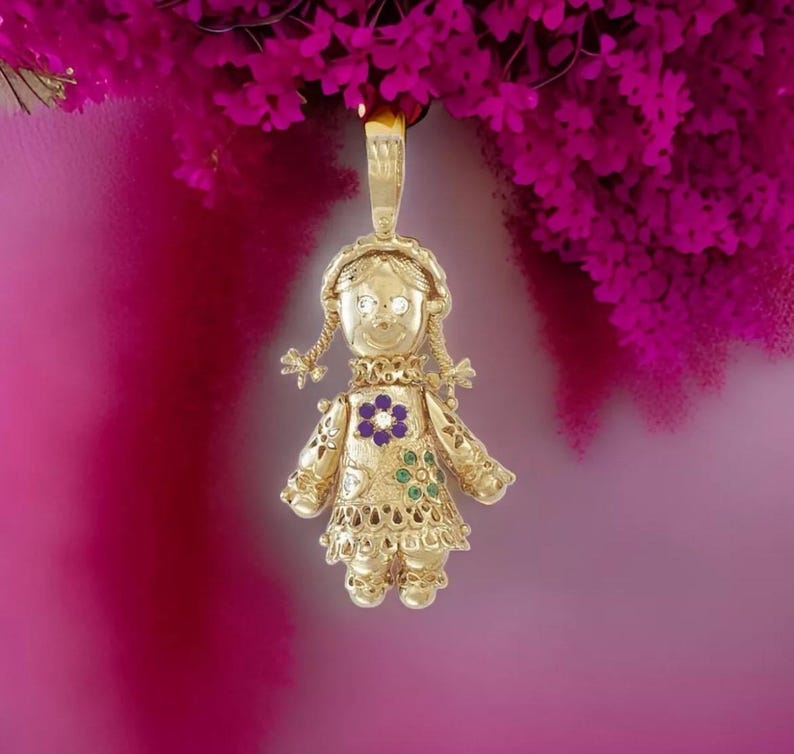 May include: A gold-colored pendant in the shape of a doll, with a smiling face, braided pigtails, and a floral design on the dress. The doll pendant has a loop at the top for a chain. The background is a gradient of pink and magenta.