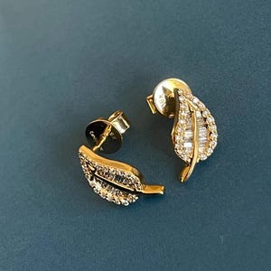 May include: A pair of gold leaf-shaped stud earrings. Each earring has a leaf design with a stem and is encrusted with small, sparkling diamonds. The earrings are set against a teal background.