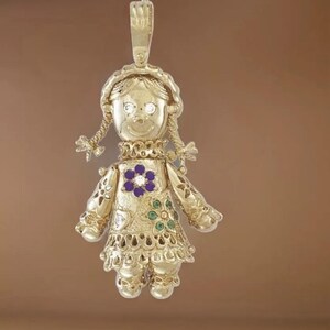 May include: A gold-colored pendant in the shape of a doll. The doll has a smiling face, braided pigtails, and a dress adorned with a purple flower and green accents. The pendant has a loop at the top for hanging.