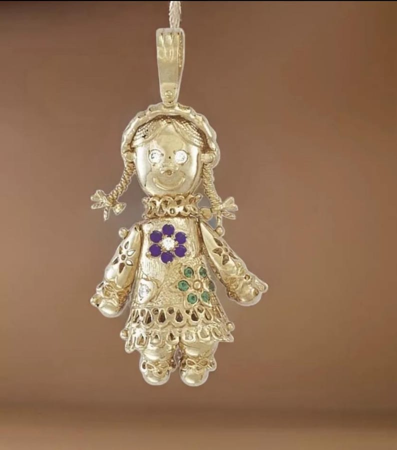 May include: A gold-colored pendant in the shape of a doll. The doll has a smiling face, braided pigtails, and a dress adorned with a purple flower and green accents. The pendant has a loop at the top for hanging.