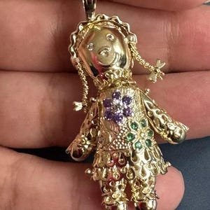 May include: A gold-colored doll pendant with a smiling face, braided pigtails, and a floral design on the dress. The pendant is adorned with small gemstones in purple and green. The doll's dress has a scalloped hemline.