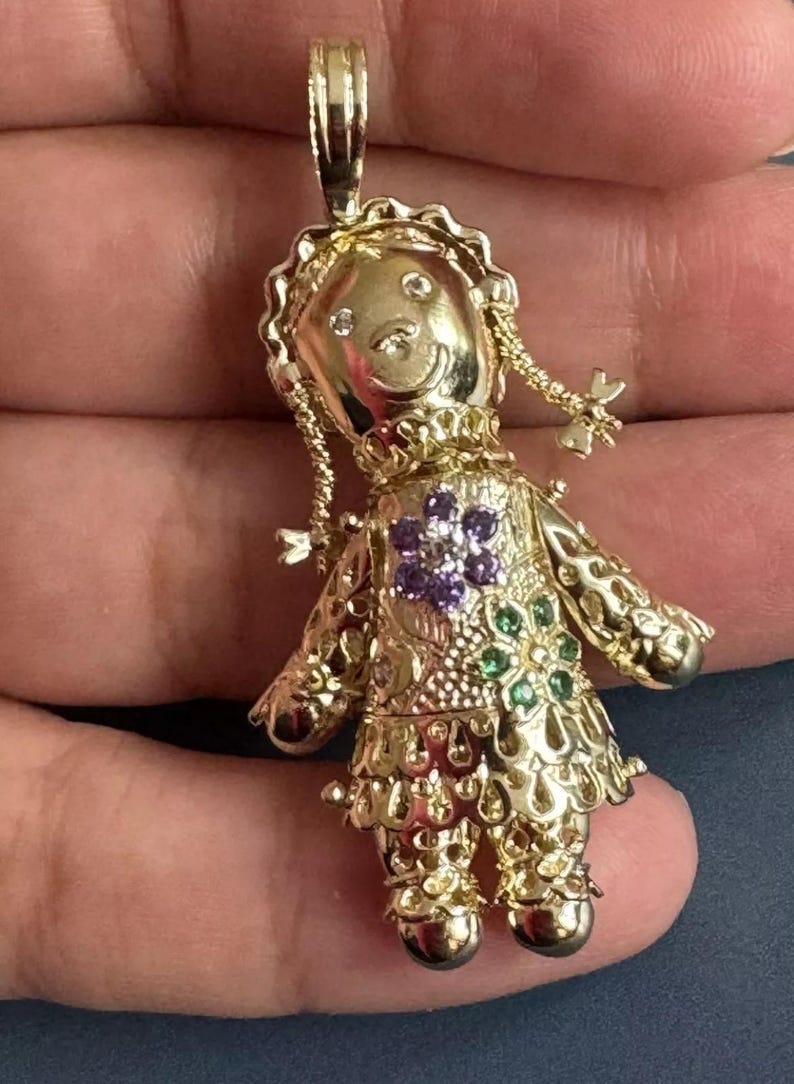 May include: A gold-colored doll pendant with a smiling face, braided pigtails, and a floral design on the dress. The pendant is adorned with small gemstones in purple and green. The doll's dress has a scalloped hemline.