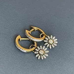 May include: A pair of gold hoop earrings with dangling diamond flower charms. The earrings are set with small diamonds and have a polished finish.