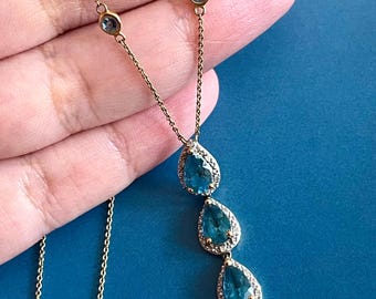 Op de afbeelding: Een goudkleurige ketting met een fijne ketting. Het heeft drie druppelvormige, lichtblauwe edelstenen, elk met een halo van kleinere, heldere stenen. Twee kleinere, ronde blauwe edelstenen zijn langs de ketting geplaatst. De ketting wordt getoond tegen een blauwe achtergrond.