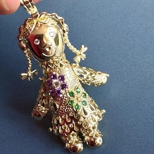 May include: A gold-colored pendant in the shape of a doll. The doll has a smiling face with two clear gemstones for eyes. The doll's dress is adorned with purple and green gemstones. The pendant is held by a hand against a blue background.