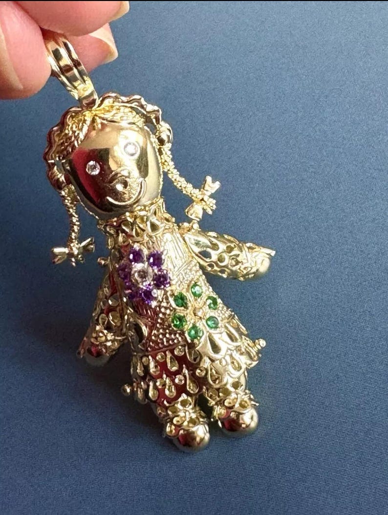 May include: A gold-colored pendant in the shape of a doll. The doll has a smiling face with two clear gemstones for eyes. The doll's dress is adorned with purple and green gemstones. The pendant is held by a hand against a blue background.