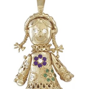 May include: A gold-toned pendant in the shape of a doll. The doll has braided pigtails, a ruffled collar, and a dress adorned with a purple flower and green accents. The doll's eyes are represented by clear gemstones.