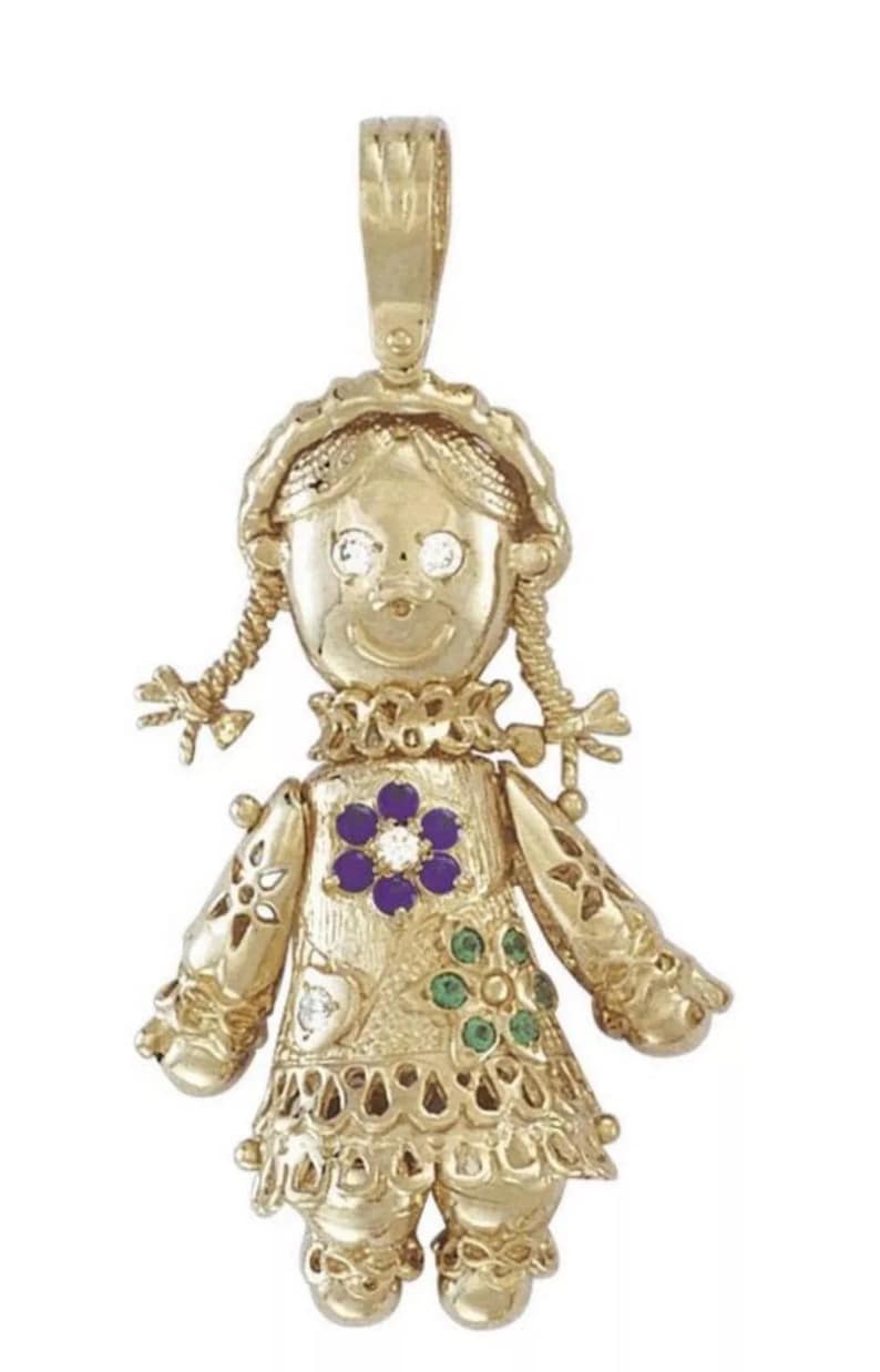 May include: A gold-toned pendant in the shape of a doll. The doll has braided pigtails, a ruffled collar, and a dress adorned with a purple flower and green accents. The doll's eyes are represented by clear gemstones.