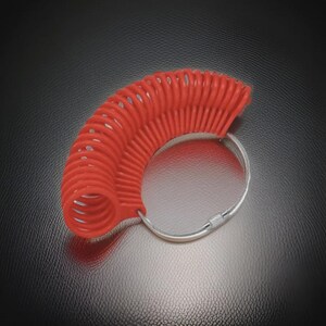 May include: A red ring sizer tool with a coiled design. The tool is made of a flexible material and is attached to a silver-colored metal ring. The ring sizer is used to measure finger sizes for rings.