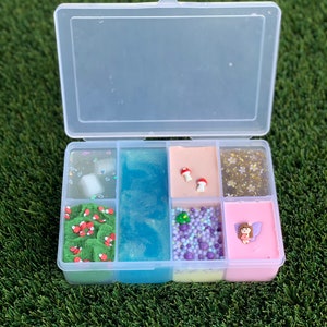 May include: A clear plastic container with six compartments filled with different colored slime. Each compartment has various embellishments, including glitter, beads, and miniature mushrooms.