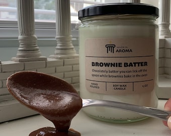 May include: A glass jar candle with a black lid and a label that reads "BROWNIE BATTER." A spoonful of chocolate batter is held in front of the candle. The candle is 15 oz and made of soy wax.