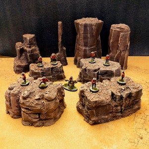 Puede incluir: Un conjunto de figuras en miniatura con uniformes rojos de pie sobre varias formaciones rocosas. Las figuras sostienen rifles y están en varias posturas. Las formaciones rocosas están pintadas en un esquema de colores marrón y gris.