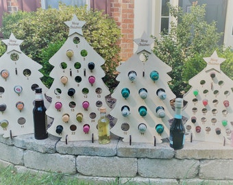 May include: Four wooden Christmas tree-shaped wine bottle holders with the text "Merry Christmas" on each. The trees are decorated with bottle caps and numbers. There are bottles of wine and champagne in front of the trees.