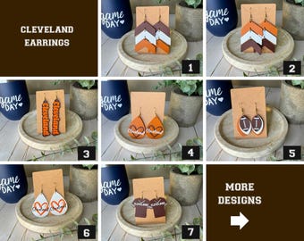 May include: A collection of seven pairs of Cleveland earrings displayed on cards. Designs include chevron patterns, football shapes, heart outlines with the word Cleveland, and jersey shapes in orange, brown, and white color schemes.