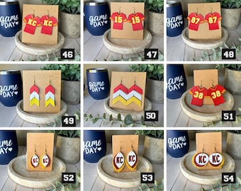 May include: A collection of nine pairs of sports-themed earrings displayed on brown cards. The designs include red jerseys with KC, 15, 87, or 38, chevron patterns in red, white, and yellow, and oval or circular shapes featuring the letters KC.