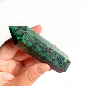 May include: A green and red gemstone point, also known as a crystal, with a faceted shape. The point is held in a hand.