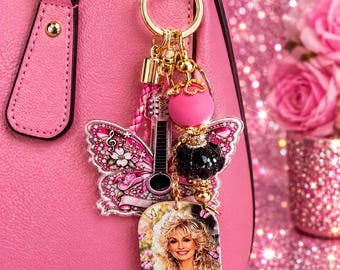 May include: A keychain featuring a gold-tone clasp, a pink butterfly-shaped charm with a guitar graphic, a large pink bead, a black faceted bead, and a rectangular charm displaying a portrait of Dolly Parton with the text R.I.P Dolly Parton.