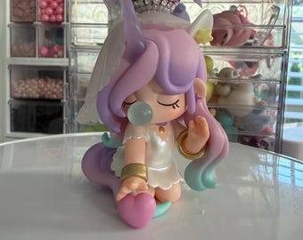 May include: A collectible figurine of a unicorn-themed character with pastel purple and teal hair, wearing a white dress and gold bracelets. The figure has a bubble in its mouth, a horn, and is holding a pink heart. It is topped with a large lavender bow.