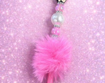 May include: A pink pen with a Betty Boop character charm, pearl and rhinestone accents, and a fluffy pink feather. The pen is set against a sparkly pink background.