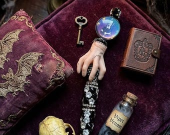 May include: A pen featuring a pale hand with stitched scars, adorned with black and clear crystals. It is accompanied by a small brown book, a metal key, a skull, and a glass bottle labeled Viper Venom containing a swirling gray liquid.