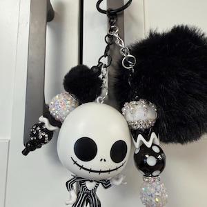 May include: A Jack Skellington keyring with a black pom-pom and beaded details. The white figure has black eyes and a stitched smile, wearing a pinstripe suit. The keyring is attached to a black ring and silver chain.