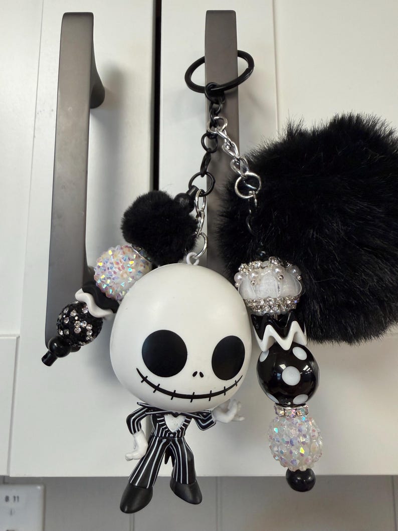May include: A Jack Skellington keyring with a black pom-pom and beaded details. The white figure has black eyes and a stitched smile, wearing a pinstripe suit. The keyring is attached to a black ring and silver chain.