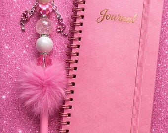 May include: A pink journal with the word "Journal" in gold lettering next to a pink pen with a fluffy feather and beaded embellishments. The pen and journal are on a pink glitter surface.