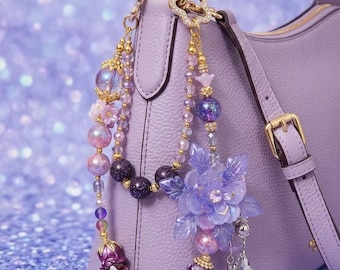 May include: A lavender pebbled leather handbag adorned with a decorative beaded charm. The charm features purple and iridescent beads, small flower-shaped accents, a large translucent purple flower, and dangling crystal-like pendants attached to a gold-tone ring.