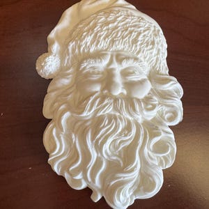 May include: A white, three-dimensional Santa Claus face sculpture. The sculpture features a detailed beard, hat, and facial features. The Santa Claus face is a festive decoration, perfect for the holiday season.