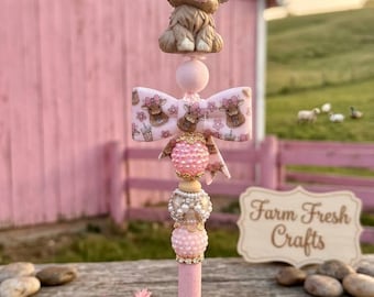 May include: A pink pen featuring a sculpted highland cow topper with a pink bow, a patterned bow bead with cow illustrations, and a stack of textured pink and white pearl-like beads with gold accents, standing on a pink base.