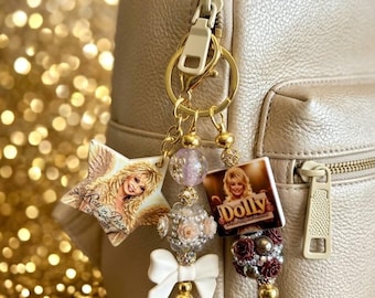 May include: A gold-toned backpack features a keychain with a star-shaped charm depicting a blonde woman with wings, a square charm labeled Dolly, a white bow, and several decorative beads in shades of pink, white, and dark red.