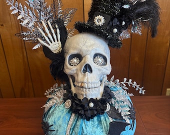 May include: A decorative Halloween pumpkin with a skull head, adorned with a black top hat, feathers, and silver accents. The teal pumpkin is embellished with a black bat, spider, and rhinestones, creating a spooky yet elegant centerpiece.