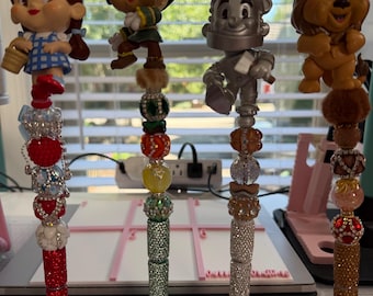 May include: Four decorative pens featuring Wizard of Oz character toppers: Dorothy, Scarecrow, Tin Man, and Cowardly Lion. Each pen has a unique stack of colorful, textured, and rhinestone-encrusted beads on the barrel, resting in a bow-shaped stand.