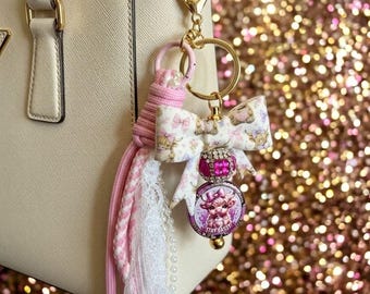 May include: A gold-toned keychain attached to a cream handbag, featuring a fabric bow with a cow print, a pink rhinestone bead, and a circular charm with a cow illustration and the text STAY SASSY, accented by pink cord and white lace.