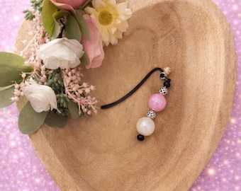 May include: A wooden heart-shaped bowl with a floral arrangement of pink roses, white and yellow flowers. A black beaded earring with pink and white beads rests in the bowl, set against a sparkly pink background.
