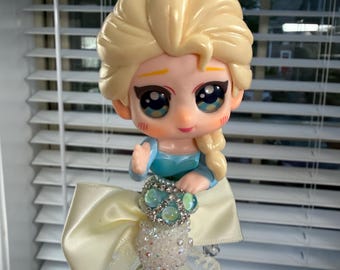 May include: A decorative figurine of a blonde princess in a blue dress, with large eyes and a sweet expression. The figurine is attached to a beaded stem with a cream-colored bow and lace accents. The beads are various colors and textures.