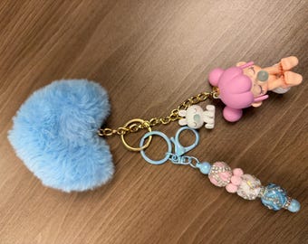 May include: A keychain featuring a light blue heart-shaped pom-pom, a pink figurine, and a beaded charm. The keychain includes a gold chain, a white bear charm, and a light blue clasp. The beaded charm has pink, white, and blue beads.