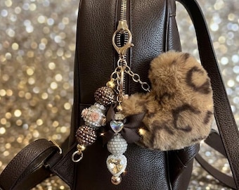 May include: A dark brown textured leather backpack featuring a decorative keychain attached to the zipper. The keychain includes a fluffy leopard print heart, a brown ribbon bow, and a strand of assorted beads in brown, white, and gold.