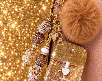 May include: A gold glitter hand sanitizer spray bottle labeled Coconut Hydrating Hand Sanitizer 1.2 fl. oz. It features a gold metal keychain with a brown faux fur pom-pom and decorative beads in shades of brown, gold, and white.
